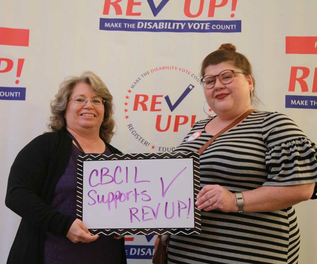 With a large REV UP banner behind them, two women smiling at the camera hold up a small dry erase board with purple writing that reads, “CBCIL supports REV UP!”
