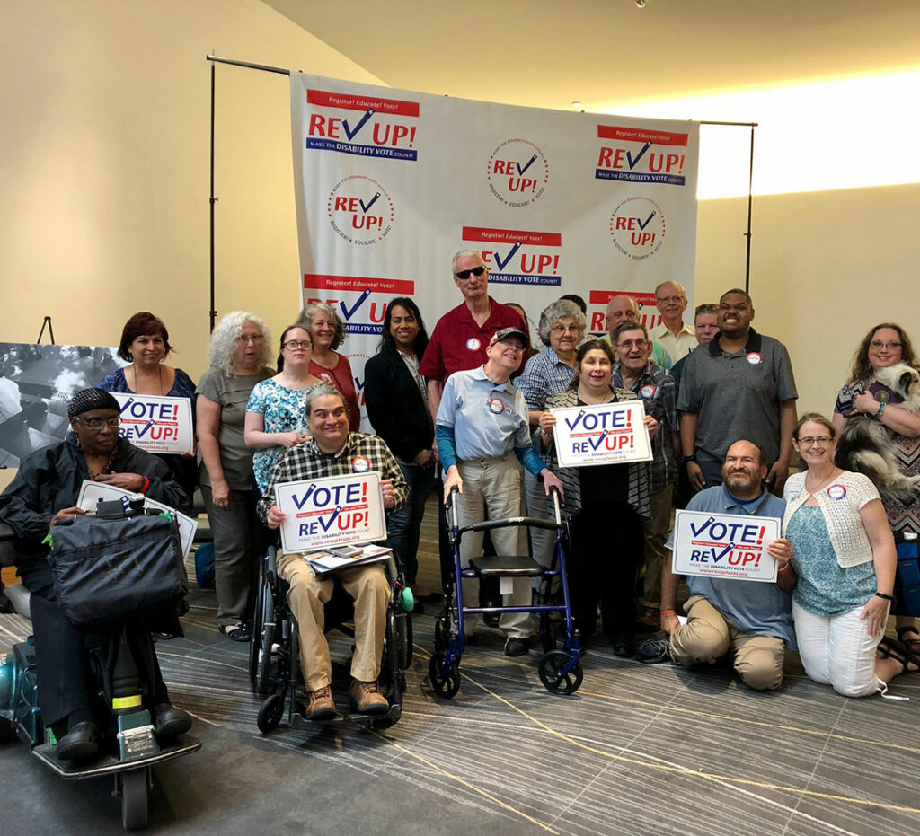Group photo of people sitting, kneeling and standing, some with assistive devices and one holding a service dog. Several hold white signs reading “Vote! REV UP!” in red and blue.
