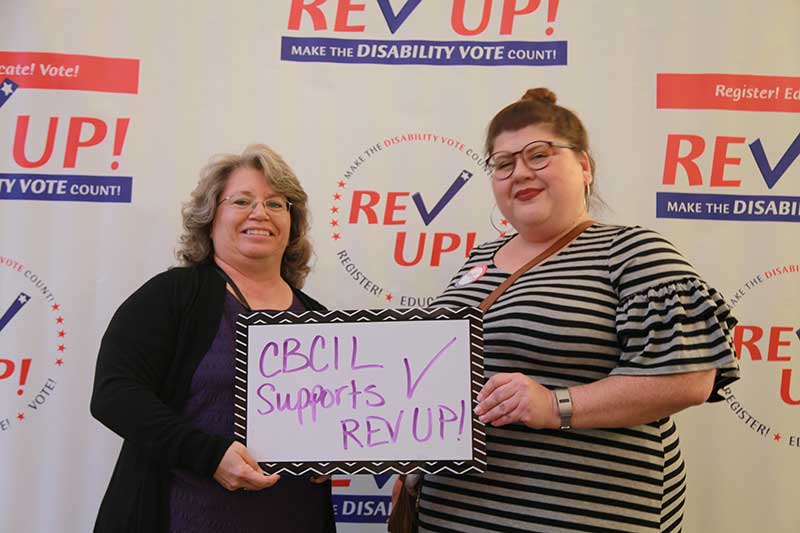 With a large REV UP banner behind them, two women smiling at the camera hold up a small dry erase board with purple writing that reads, “CBCIL supports REV UP!”