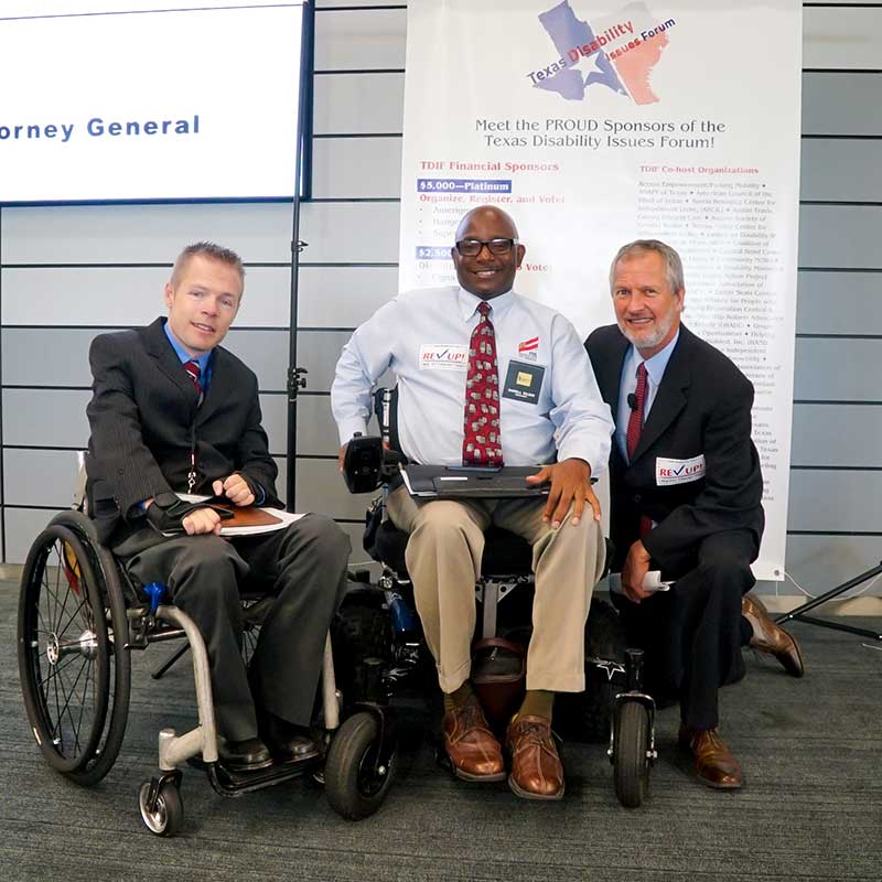 Three men in business suits, two sitting in wheelchairs and one kneeling, pose for a photo in front of a large banner listing TDIF sponsors.