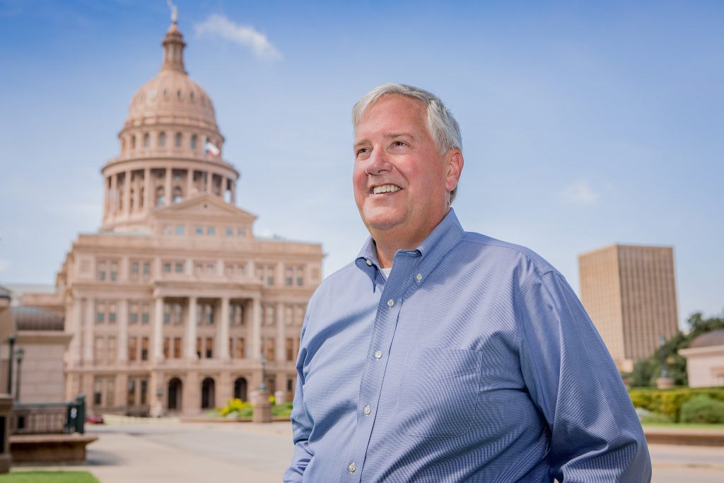 Portrait of Mike Collier from the waist up, smiling and looking into the distance. The Texas Capitol is in the background.
